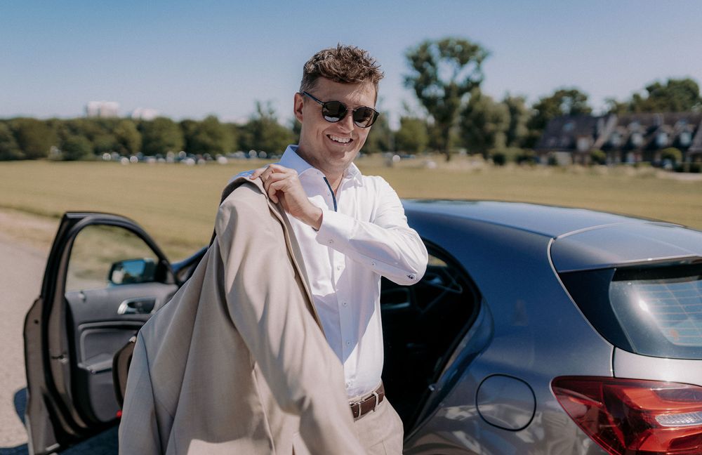 Person in white shirt and sunglasses leans against dark car on sunny day.