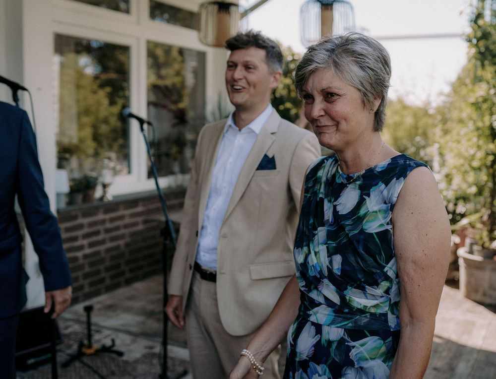 Wedding guests in formal attire smile while standing outside brick building.