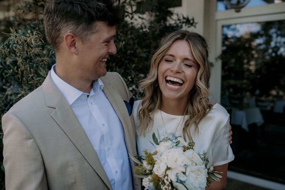 Couple laughs together while holding white floral bouquet against greenery backdrop.