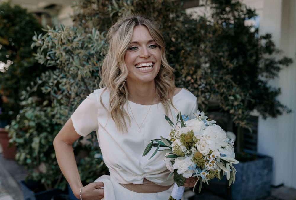 Bride in modern two-piece wedding dress holds bouquet while smiling outdoors.
