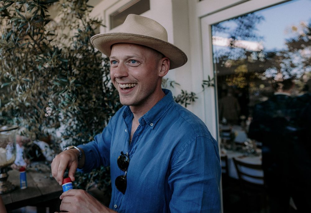 A person wearing a blue shirt and tan hat smiles while enjoying a drink outdoors.
