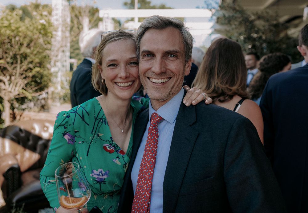 A happy couple in formal attire smiling at a garden party with drinks.