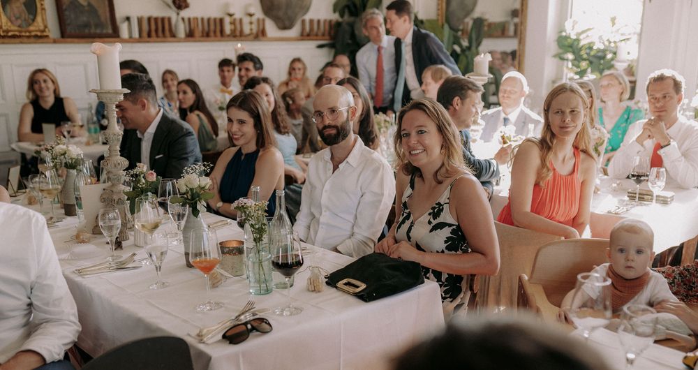 Wedding reception guests seated at long tables in a bright, window-lit venue.