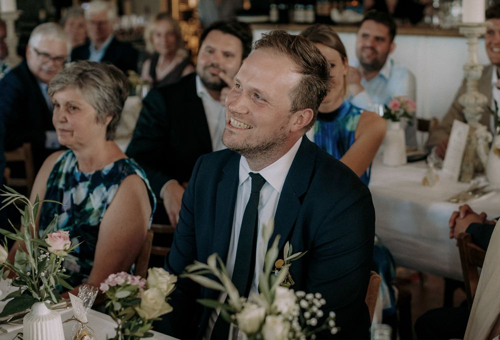 Wedding guest in a blue suit smiles while seated at decorated reception table.