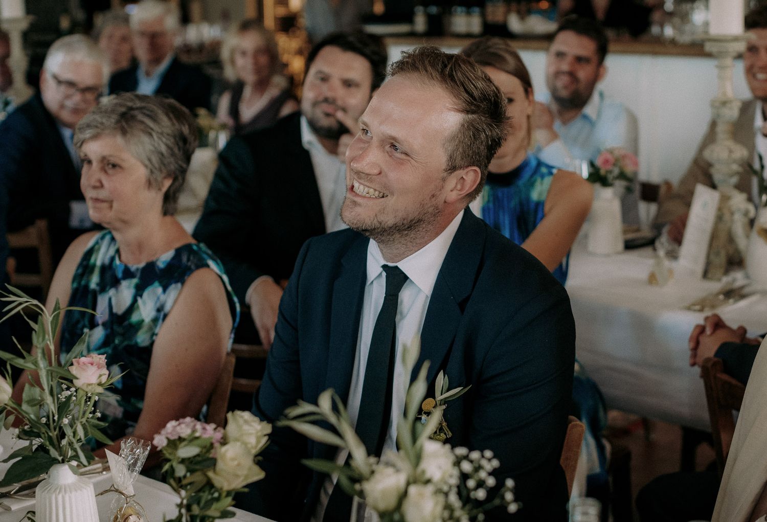 Wedding guest in a blue suit smiles while seated at decorated reception table.