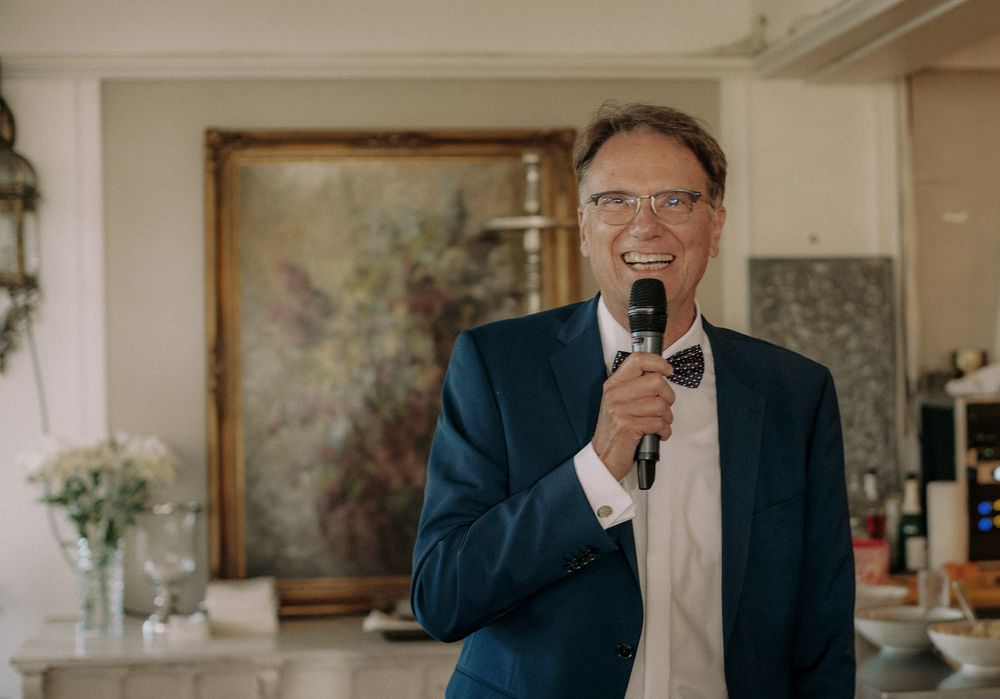 A speaker in a navy suit smiles while delivering a speech with a microphone at an indoor event.