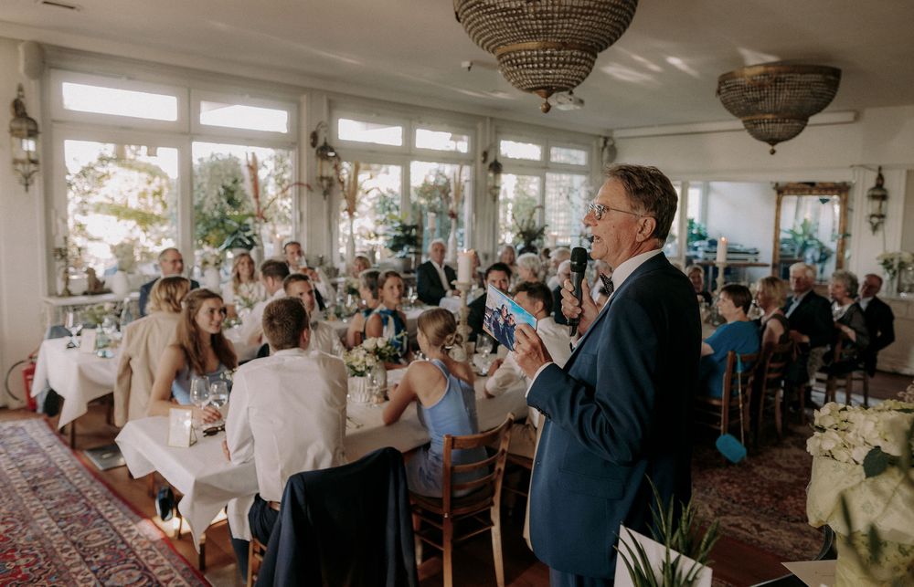 A speaker addresses a crowded dining room filled with wedding guests under elegant chandeliers.