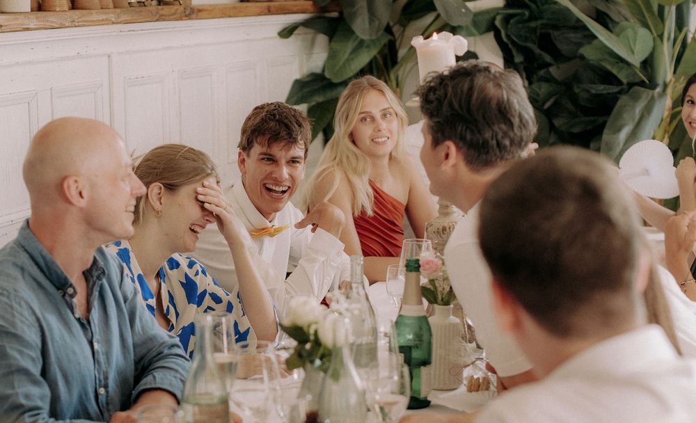 Wedding guests laugh and socialize around a dining table decorated with greenery.