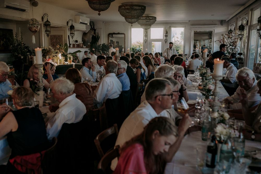 A crowded wedding reception dining room filled with guests at long tables enjoying a meal and celebration.