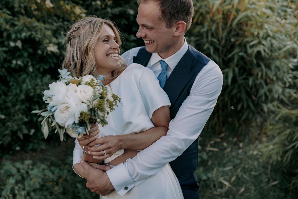 A joyful wedding couple embracing outdoors with white floral bouquet.