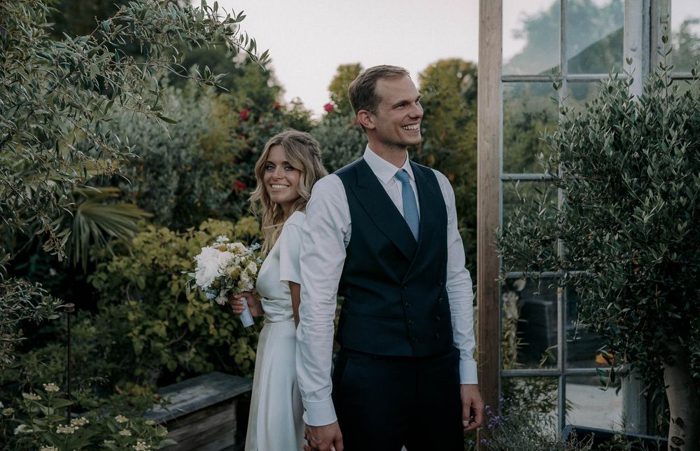Elegant wedding couple posing in front of greenhouse windows surrounded by greenery.