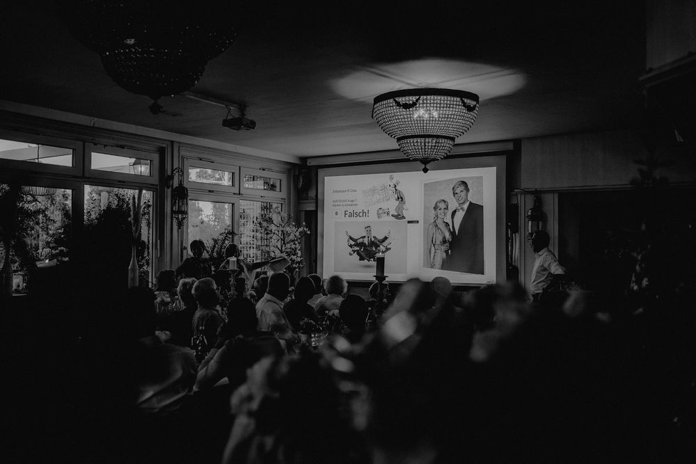 A dimly lit room filled with people watching a presentation or performance with projected images.