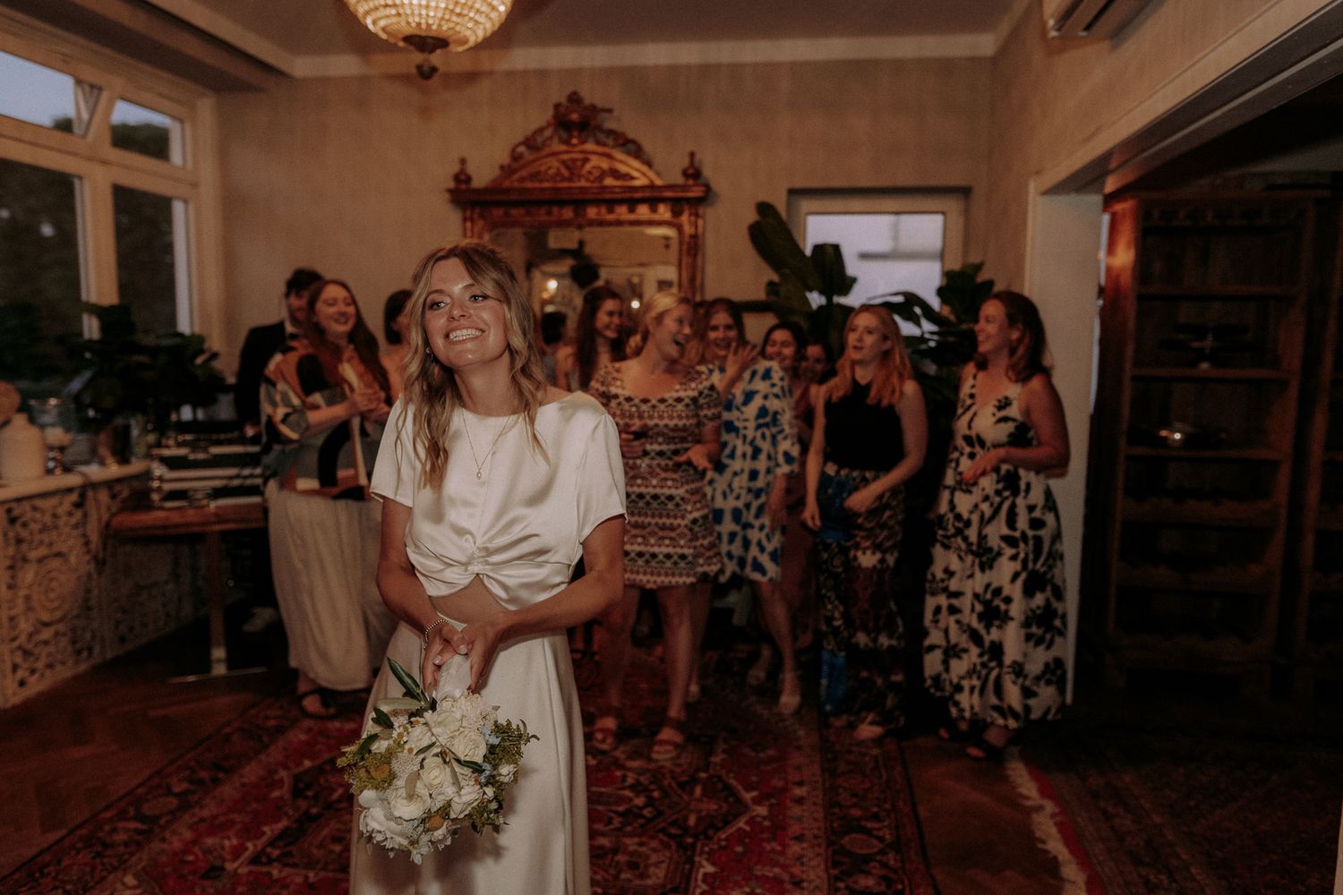 A bride in a white dress holds a bouquet while guests in floral dresses watch during a wedding reception.