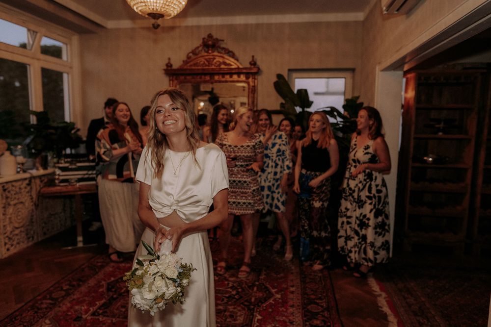 A bride in a white dress holds a bouquet while guests in floral dresses watch during a wedding reception.