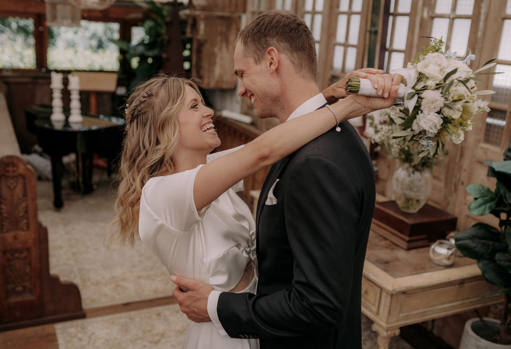 Newlyweds share an intimate embrace while holding a white floral bouquet in a rustic indoor setting.