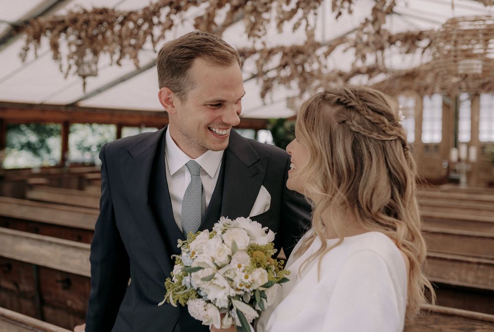 Happy couple shares joyful moment together while holding white wedding bouquet in sunlit venue.