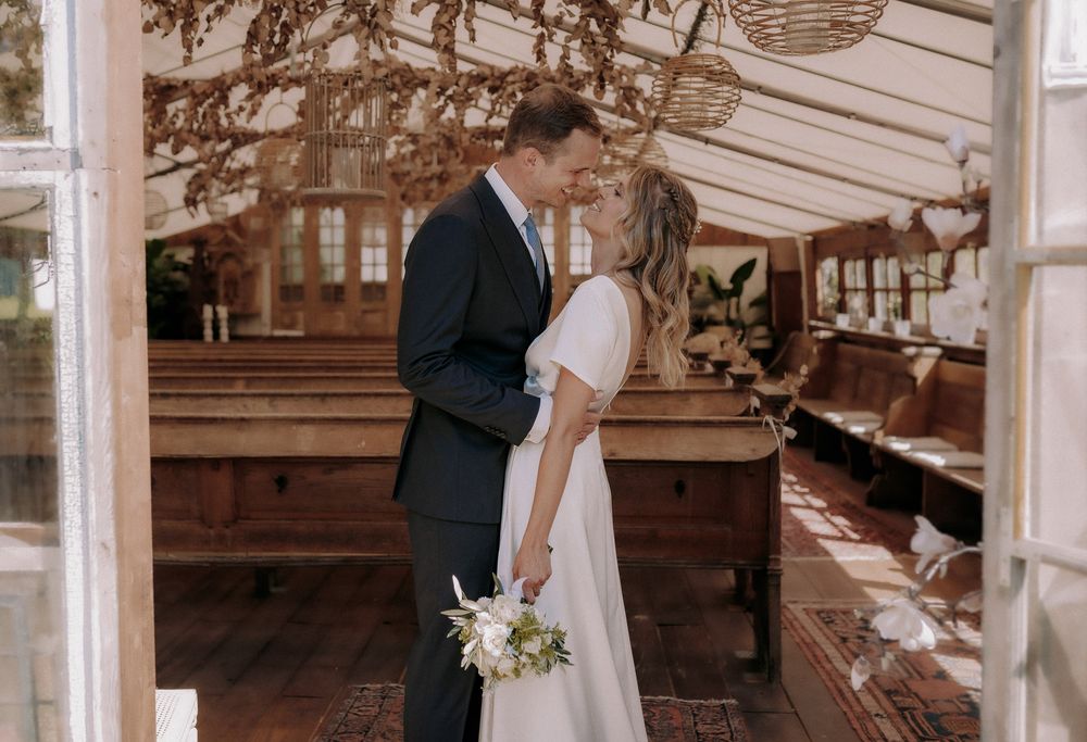 Couple in wedding attire stands together in rustic wooden venue with natural lighting.