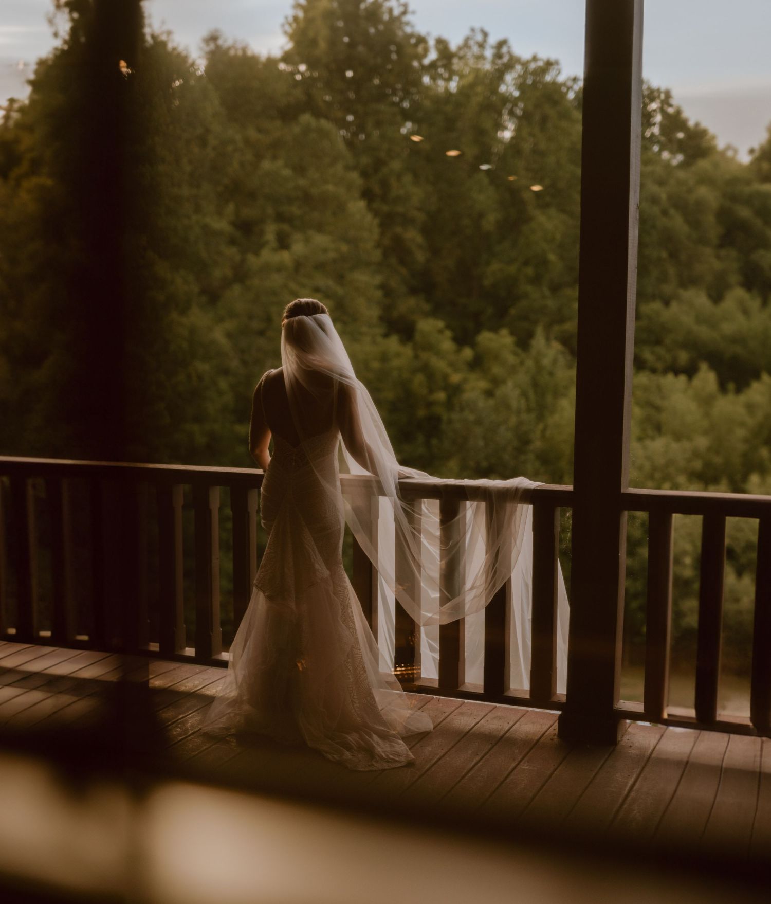Bride in lace gown with flowing veil stands on wooden deck overlooking forest view.