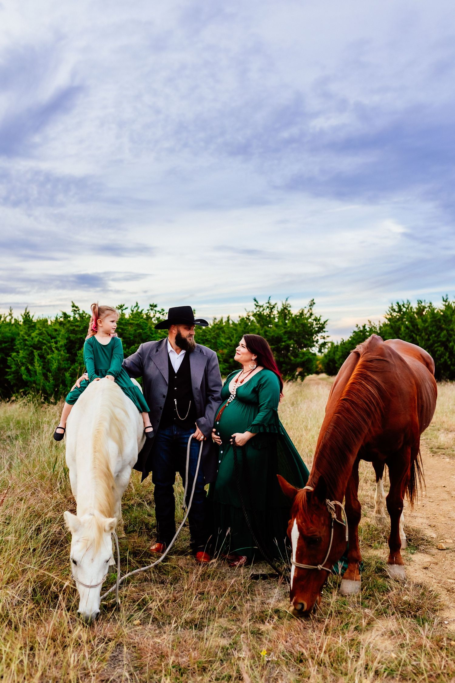 Family in vintage attire poses with two horses in a rustic field beneath cloudy skies.