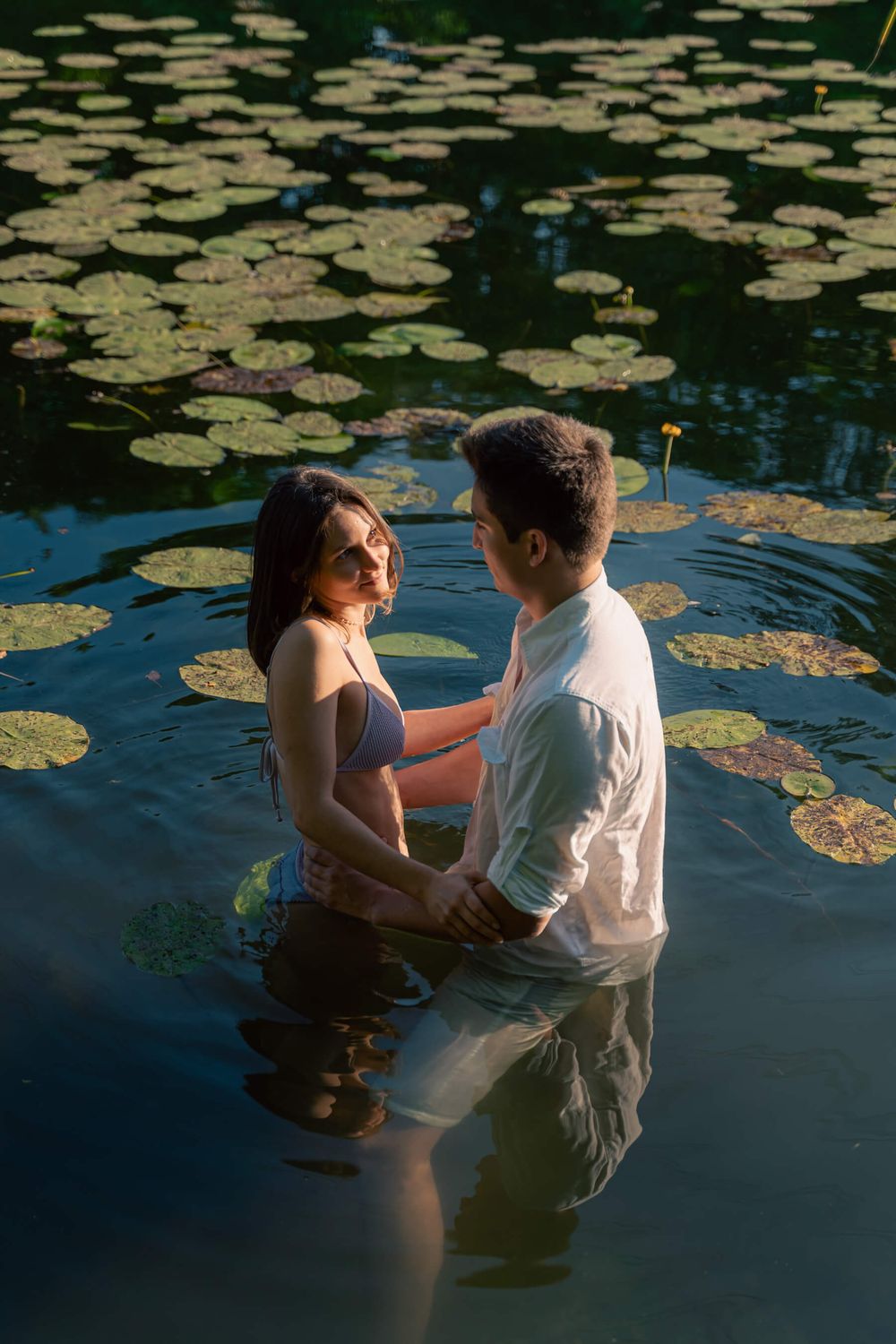 A couple stands intimately in a pond surrounded by floating lily pads at sunset.