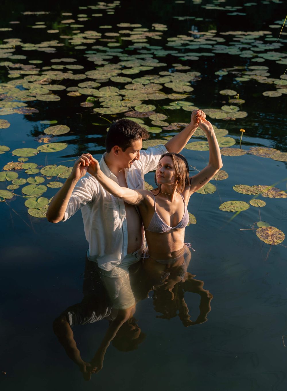A couple dances playfully in a lily pad covered pond during golden hour.