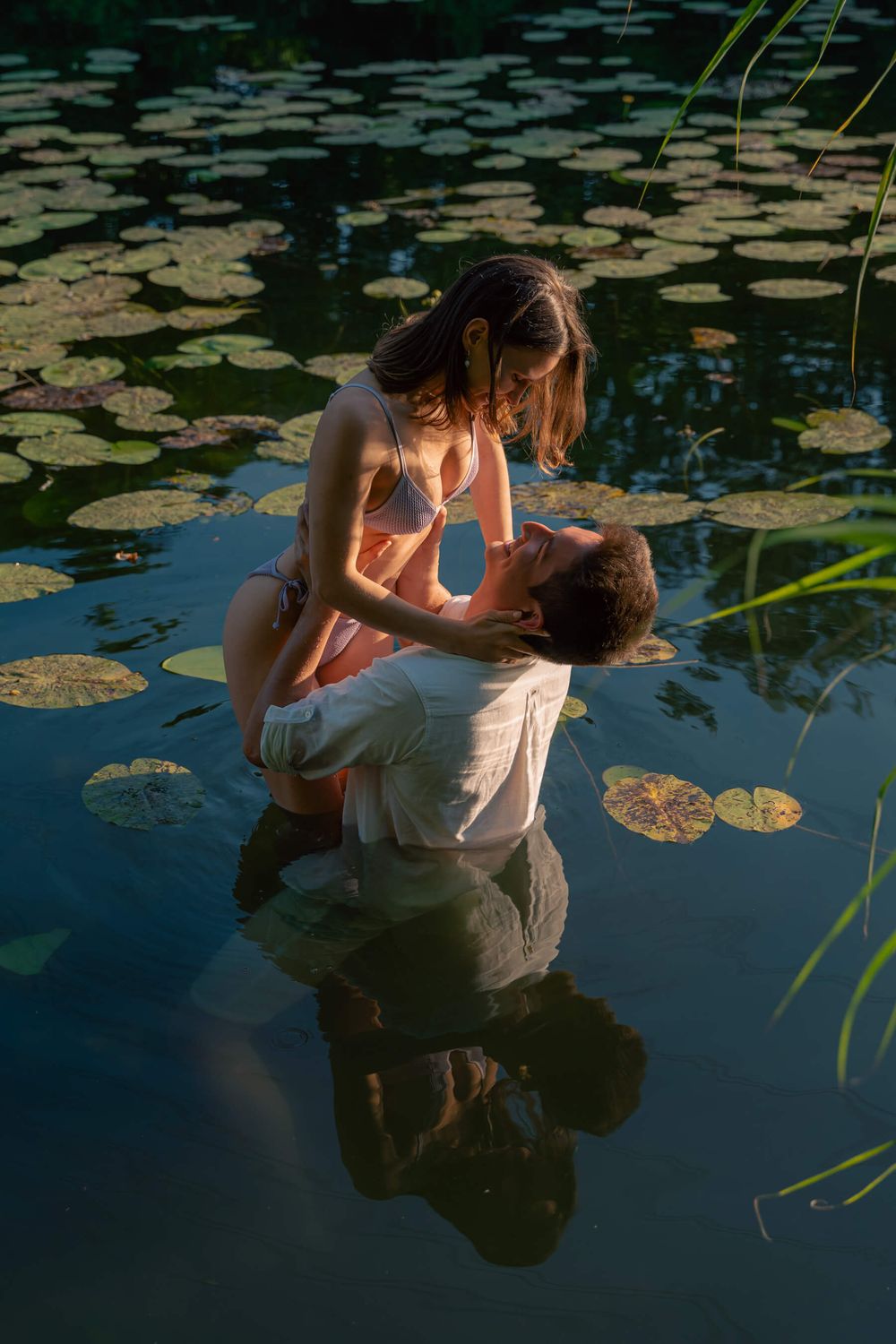 A romantic couple poses in a pond surrounded by lily pads during sunset.