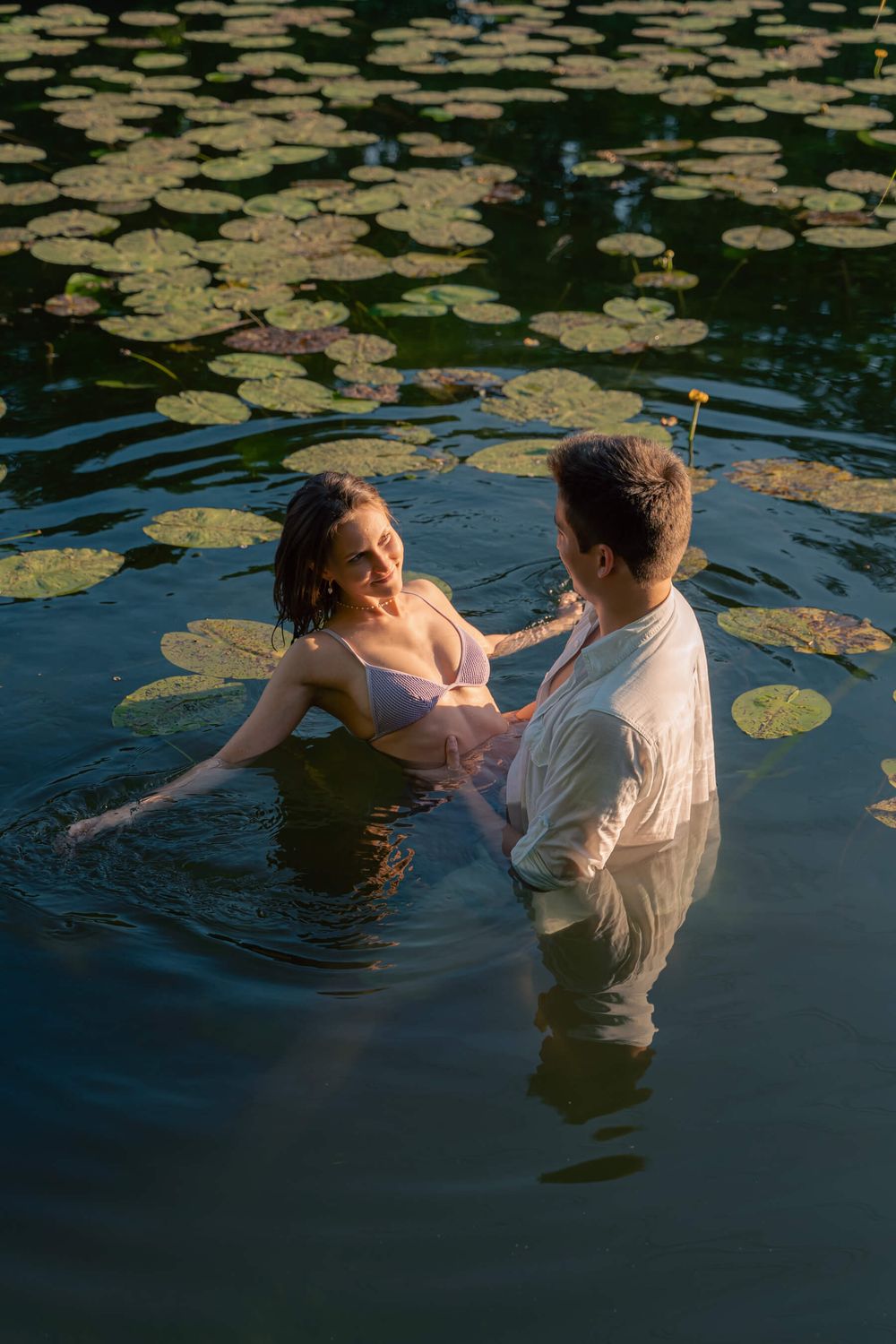 Romantic couple embraces in a lily pond during sunset, surrounded by floating lily pads on dark water.
