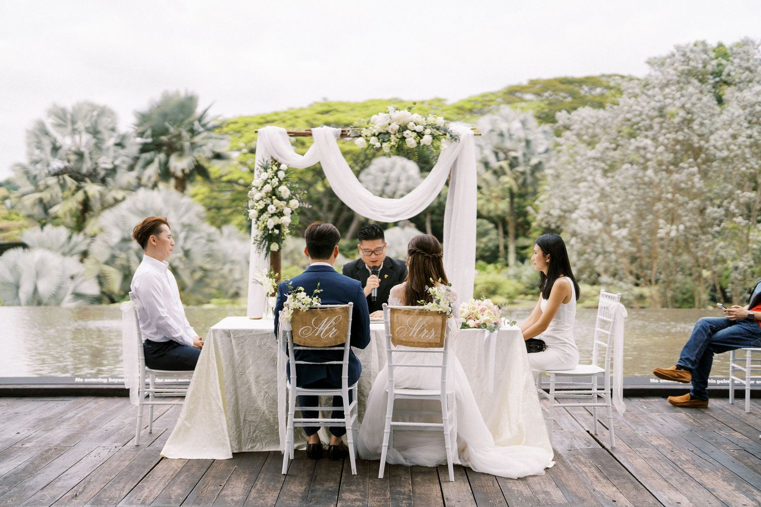 Wedding ceremony setup on wooden deck with white arch and chairs overlooking scenic landscape.