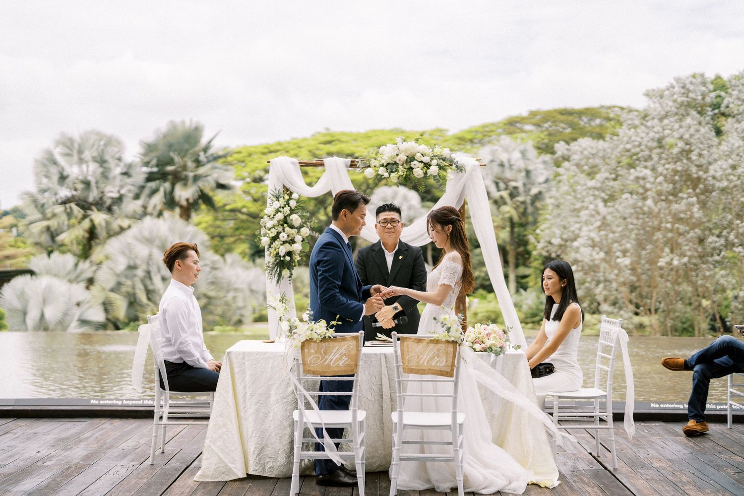 Intimate outdoor wedding ceremony with white draped arch and chairs on waterfront deck.