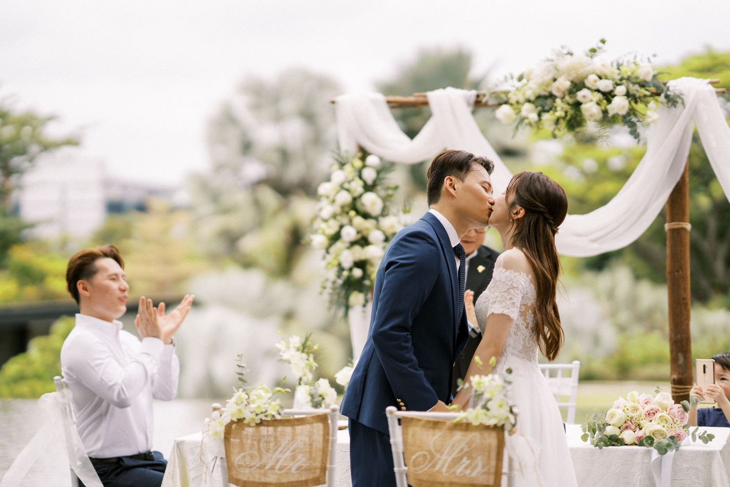 Garden wedding ceremony setup with white floral arch and wooden chairs under natural lighting.