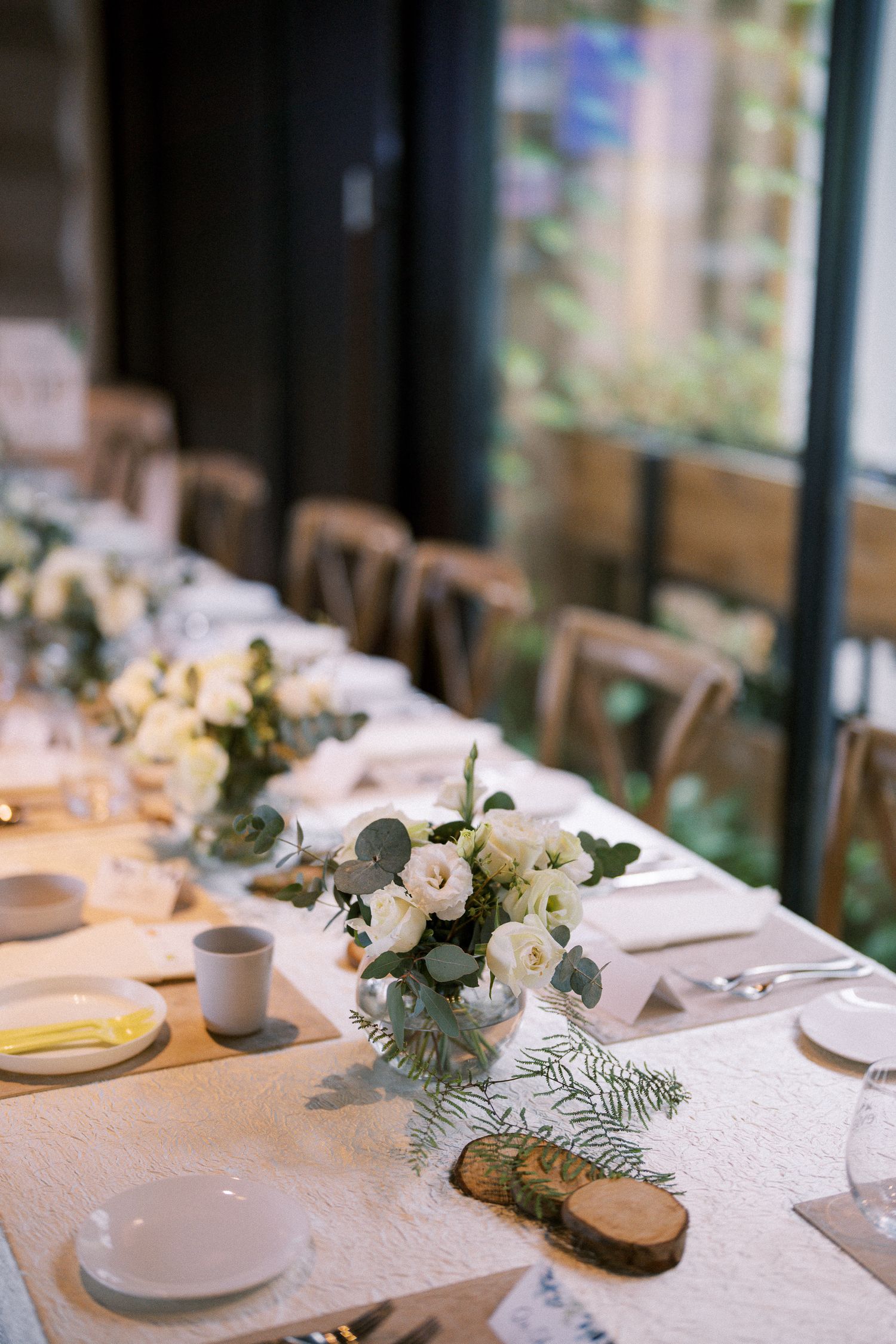 Elegant white wedding reception tables with floral centerpieces and natural light from large windows.