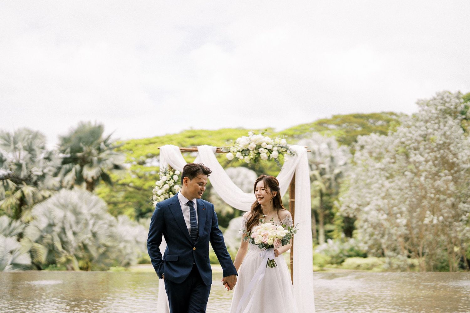 Newlyweds walking together under white floral arch with scenic water and hillside backdrop.