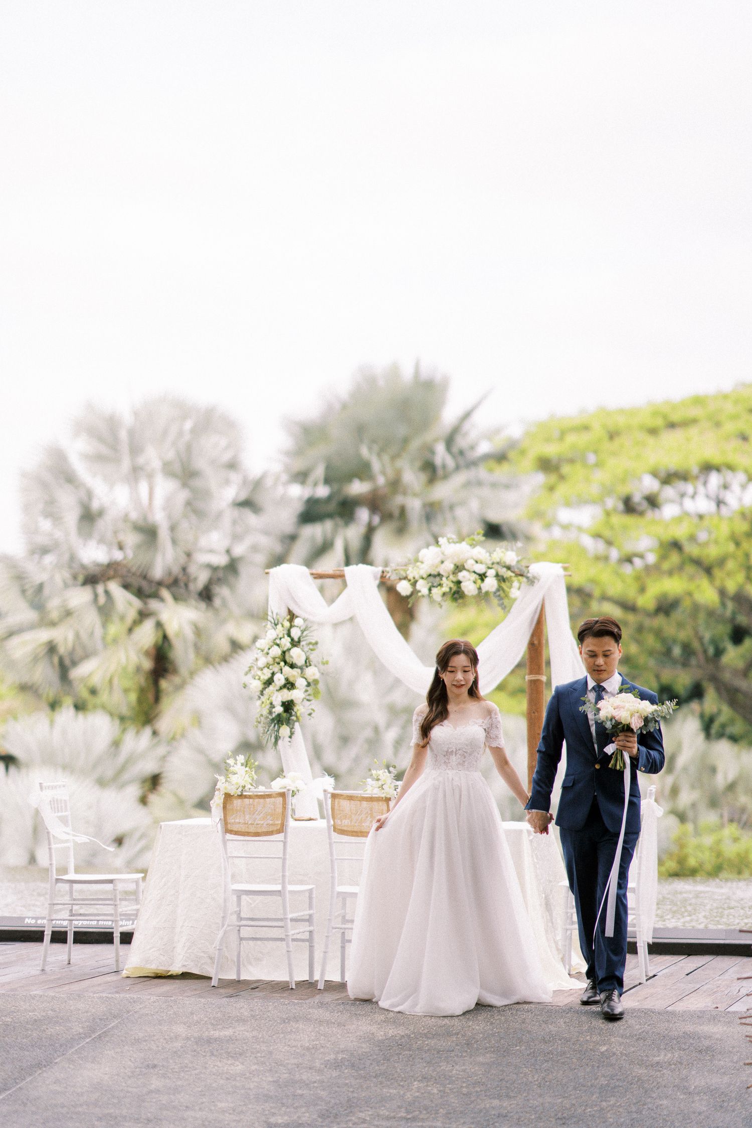 A couple stands under an elegant white wedding arch with floral decorations and white chairs in an outdoor garden setting.