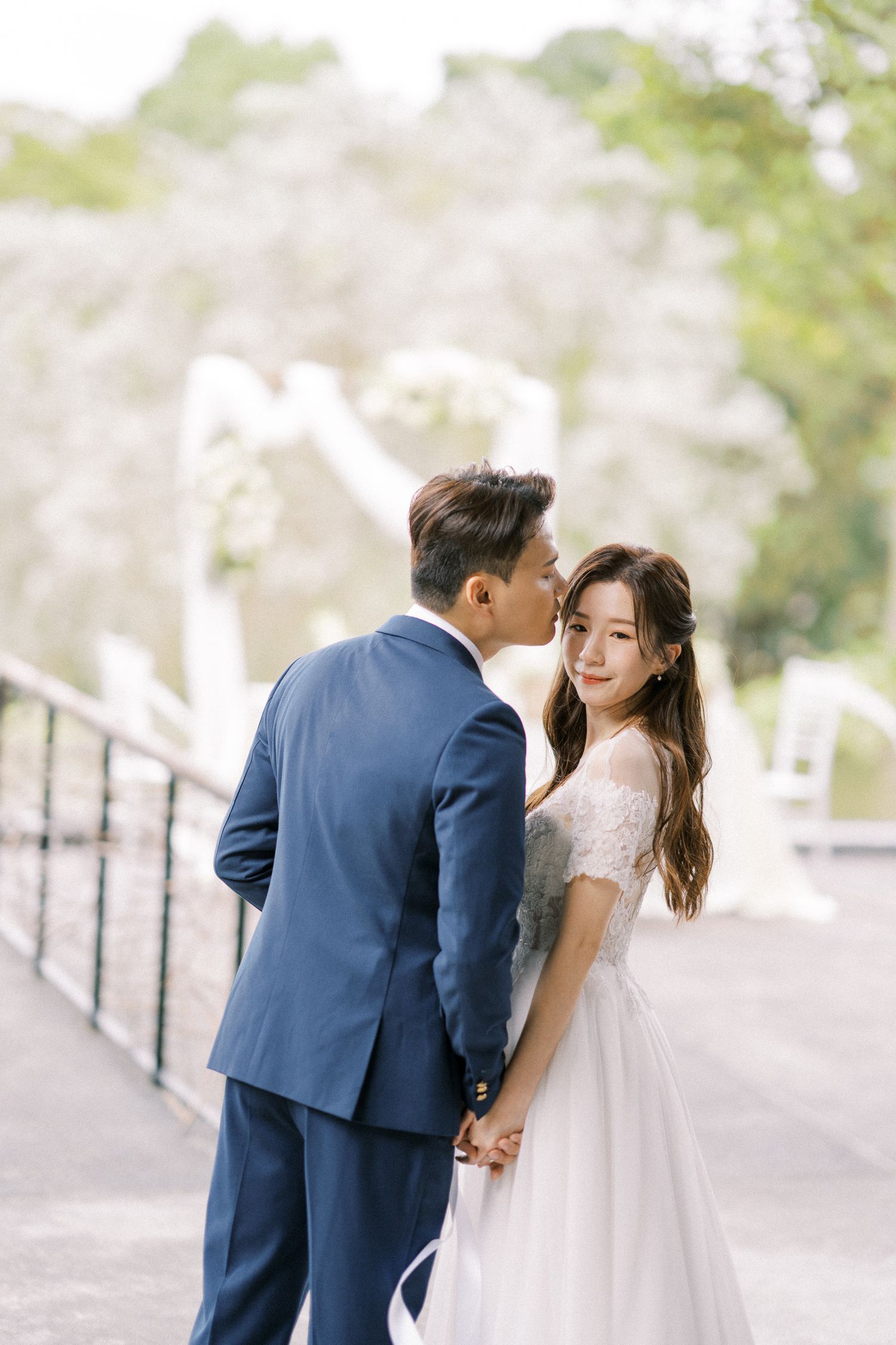 A romantic wedding moment captured on a bridge with soft natural lighting and lush greenery in the background.