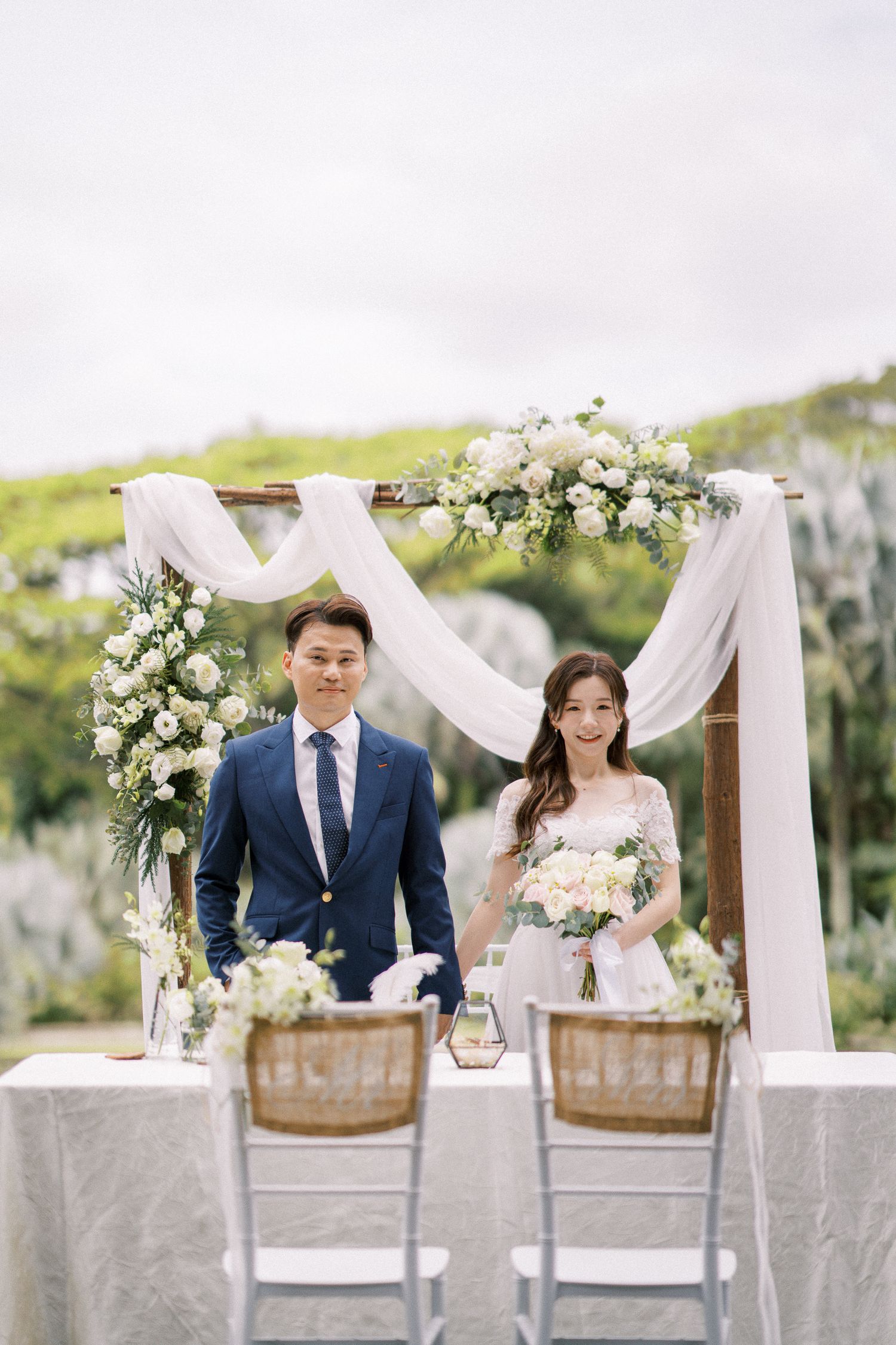 An intimate wedding setup with white draping, floral arch and rustic chairs against a scenic natural backdrop.
