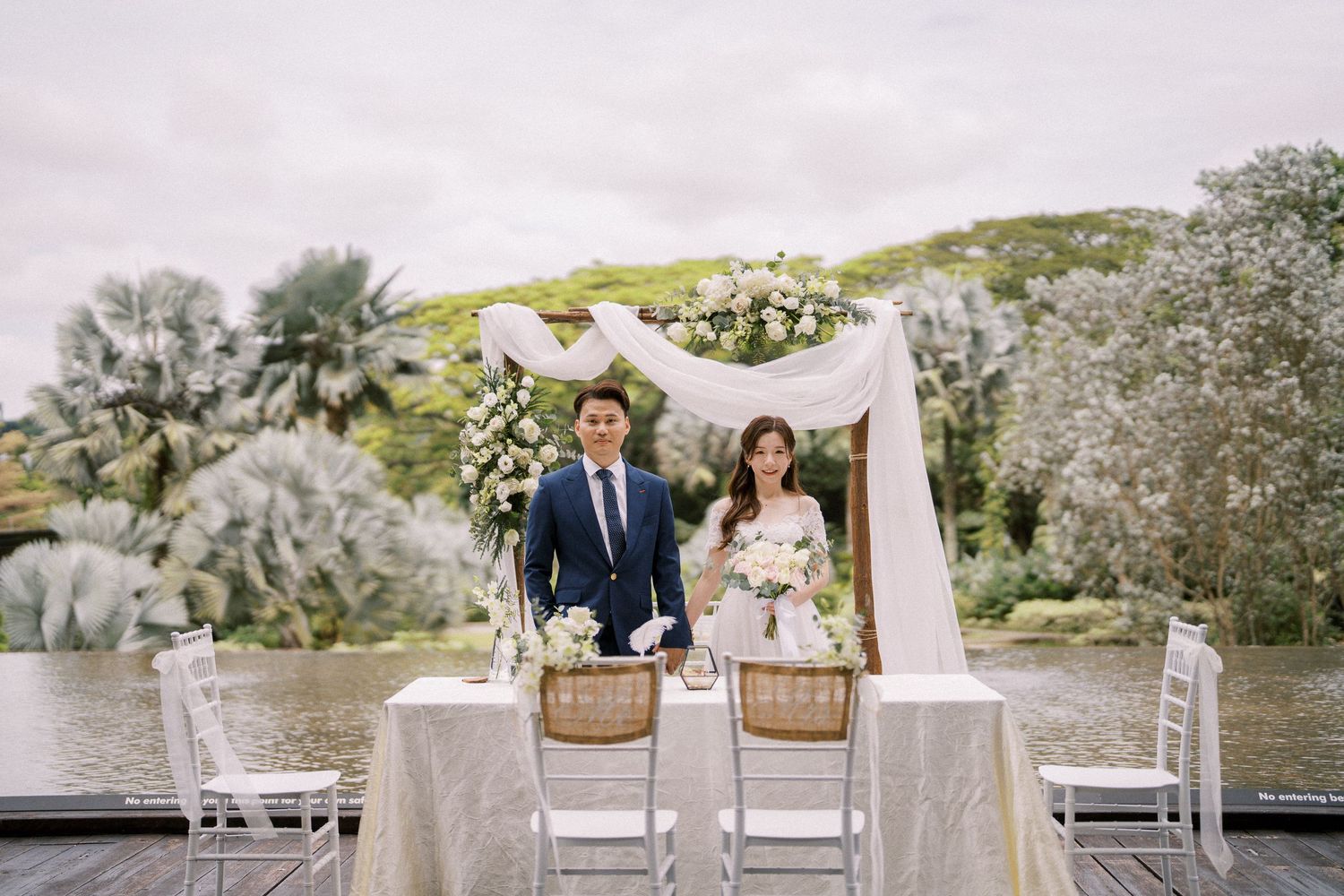 A wedding ceremony setup on a wooden deck overlooking water with white draping and floral decorations.