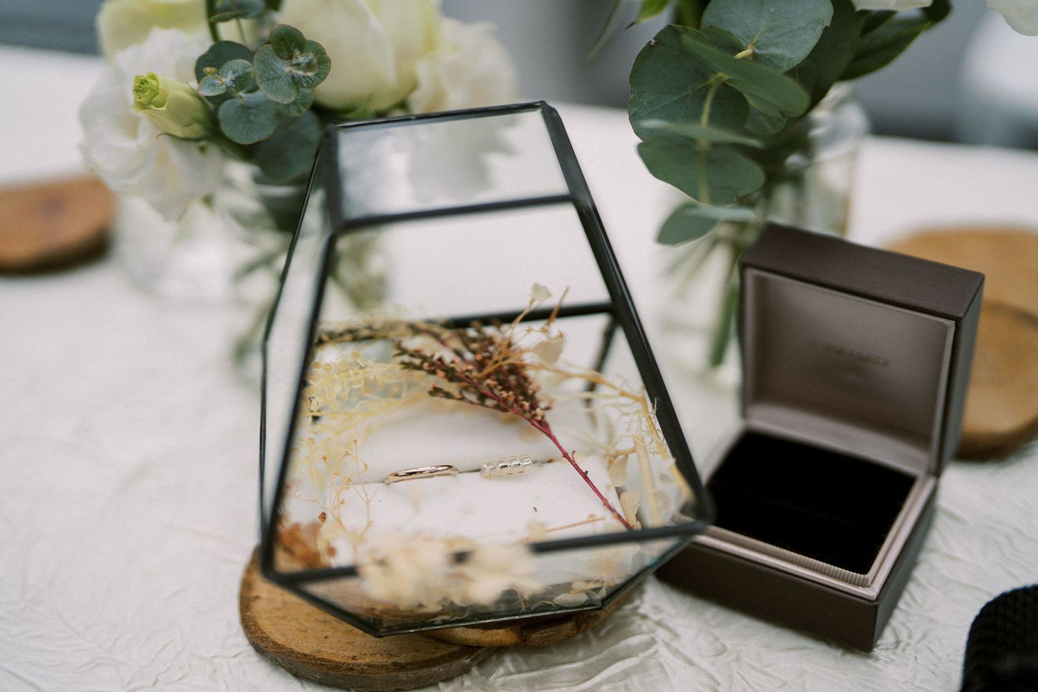 Wedding details with geometric terrarium and ring box on white tablecloth with floral arrangements.