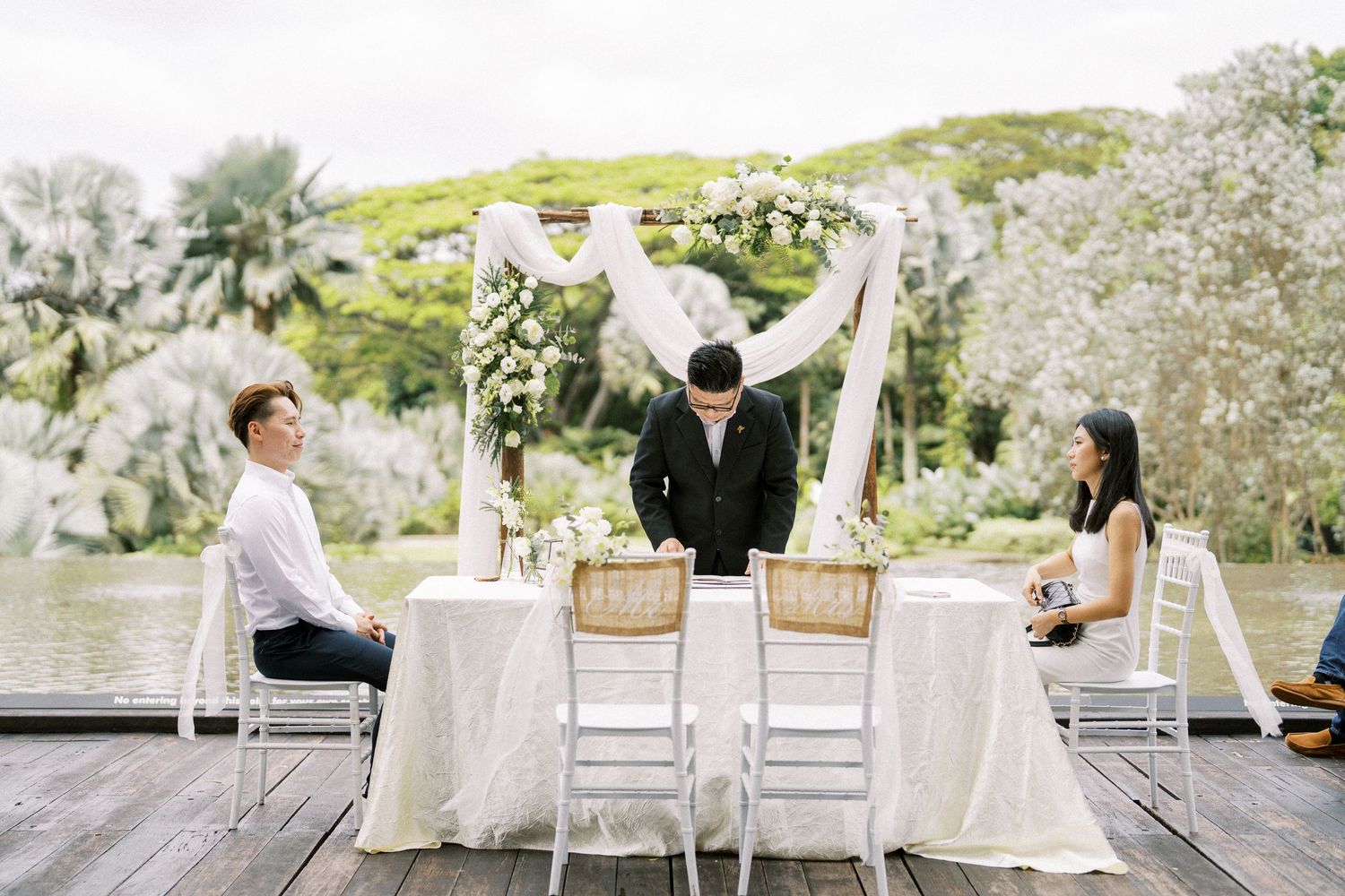 A white outdoor wedding setup with chairs and floral arrangements against a natural backdrop.