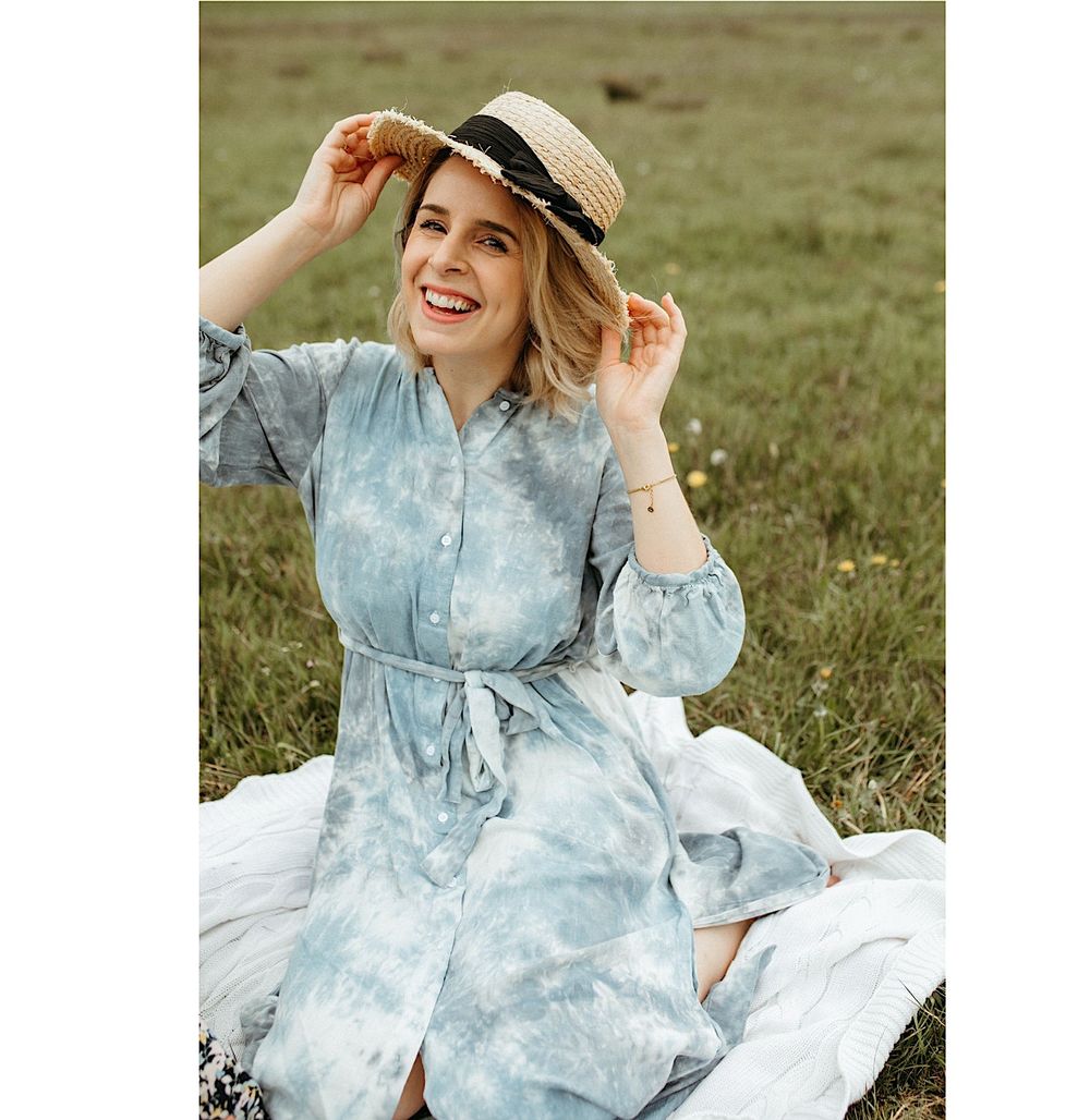 Woman in blue tie-dye dress poses on white blanket in sunny meadow.