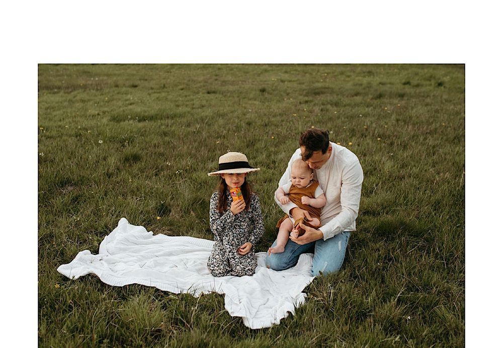 A couple sits together on a white blanket in a grassy field during a cloudy day.