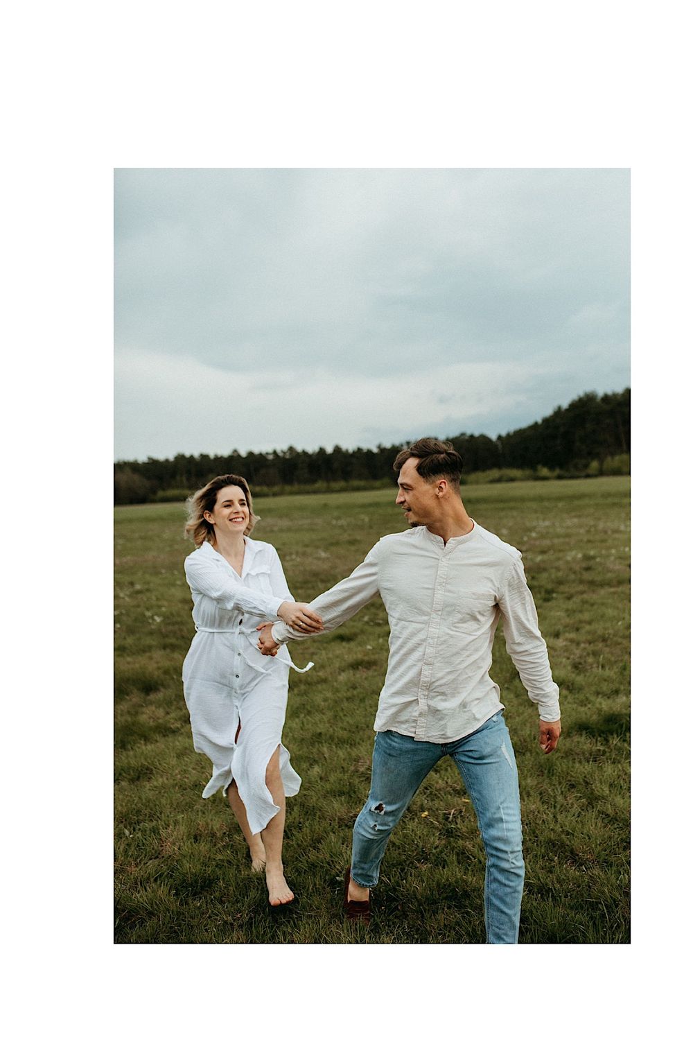 A playful couple in white shirts runs through an open field on a cloudy day.