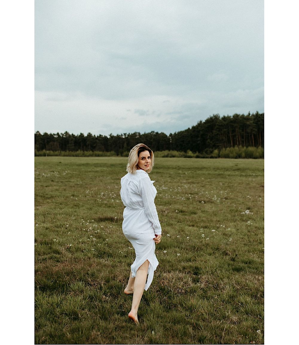 A figure in a flowing white dress dances freely in a vast meadow landscape.