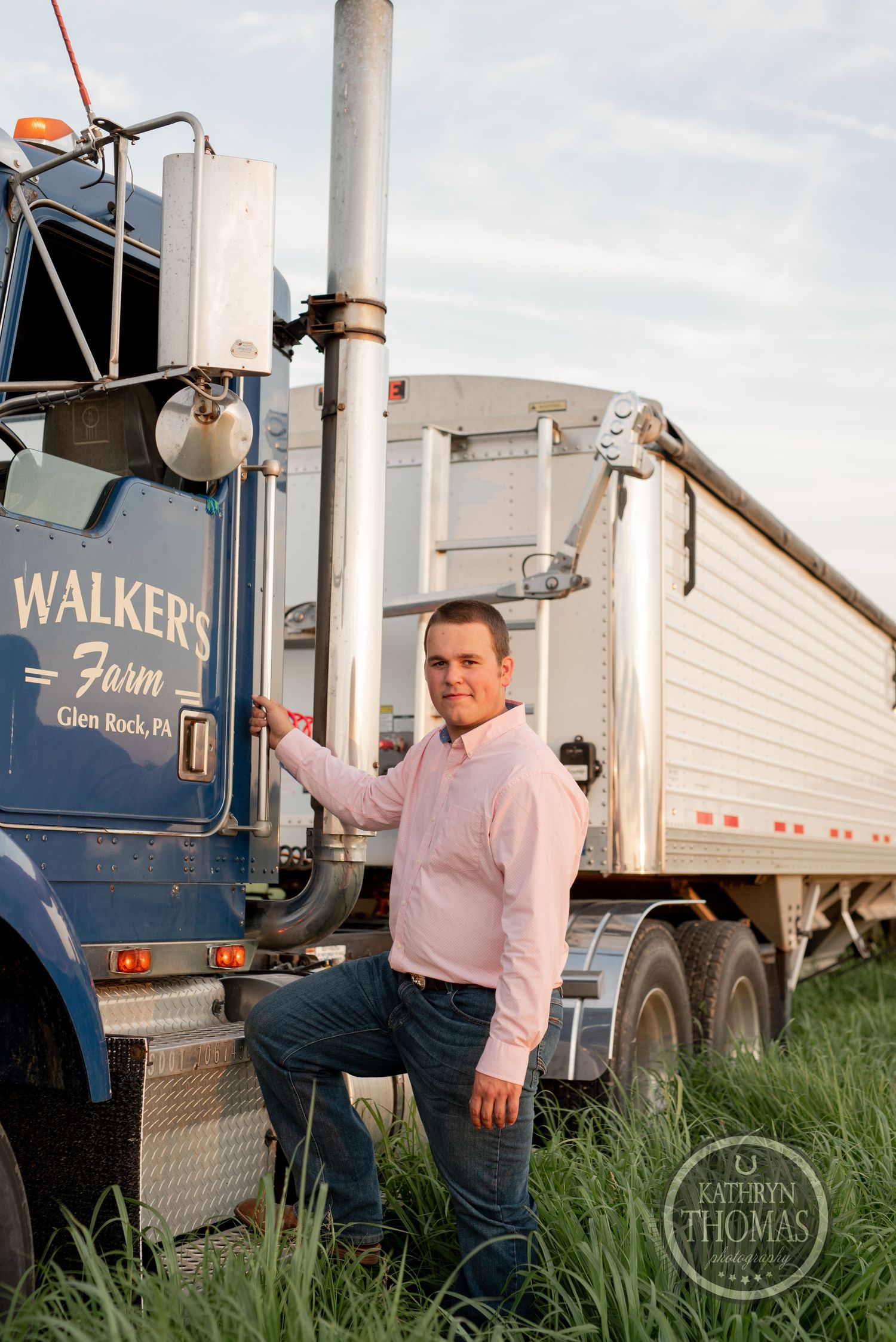 Driver poses beside Walker's Farm branded blue semi truck in grassy field.