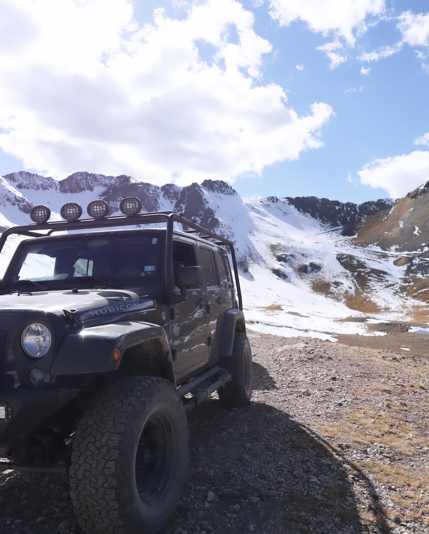Black Jeep Wrangler parked on rocky mountain terrain with snowy peaks and blue skies in background.