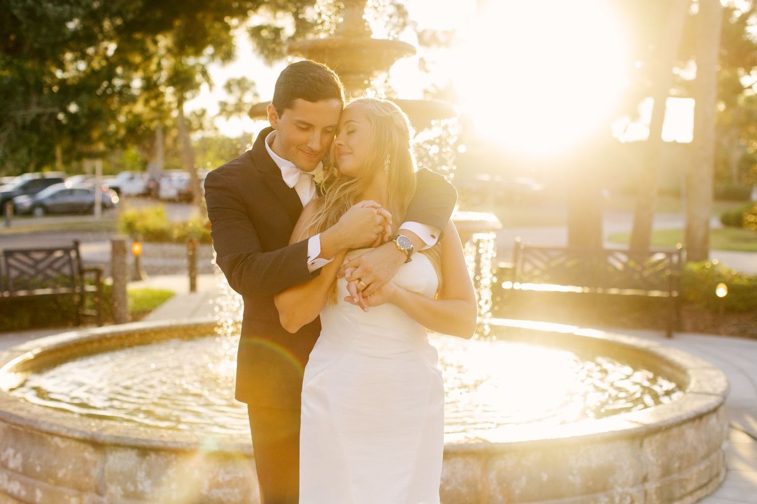 A romantic sunset wedding photo of a couple embracing by a fountain.