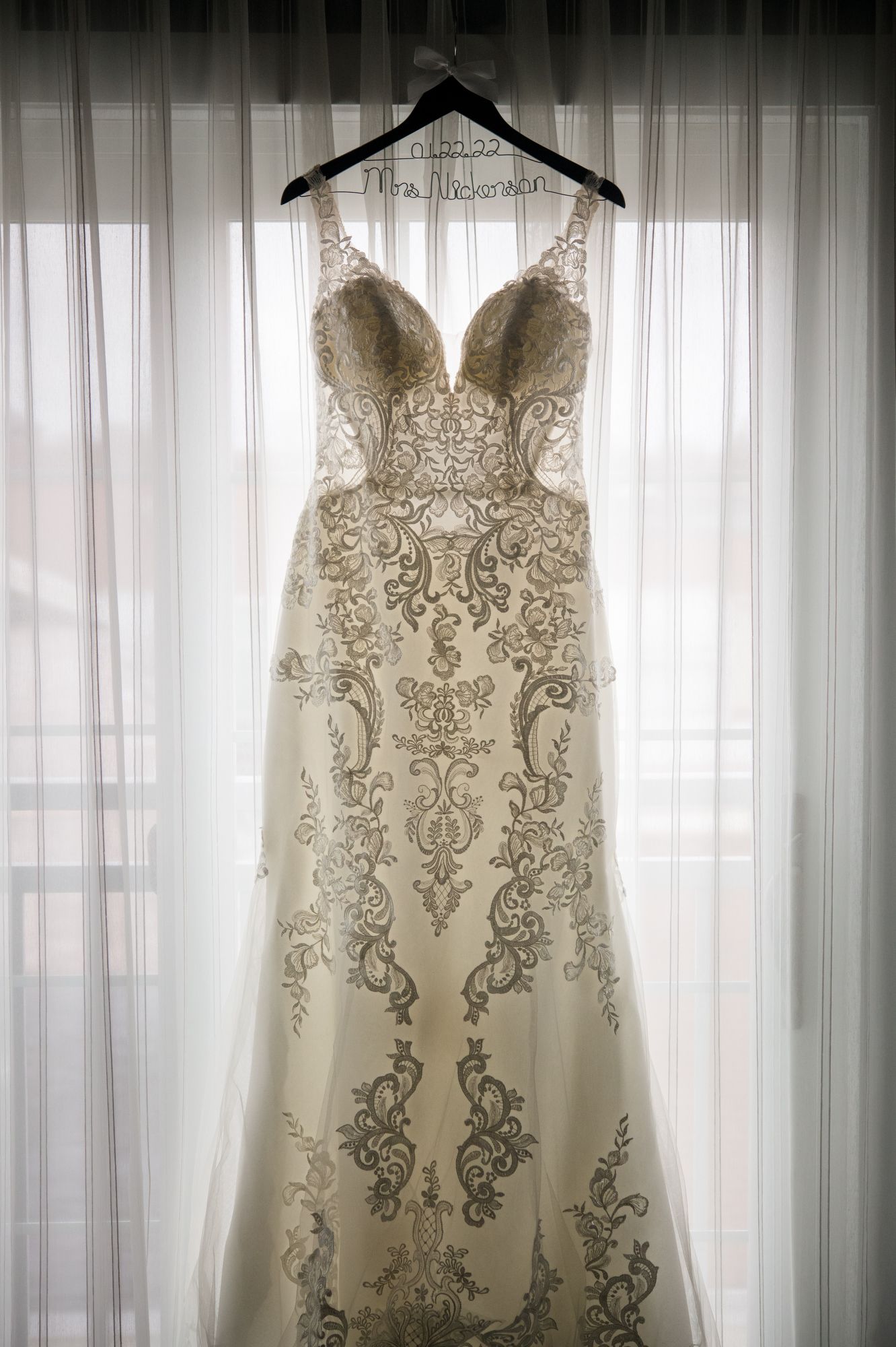 Elegant lace wedding dress with intricate beading hangs in window light against sheer curtains.