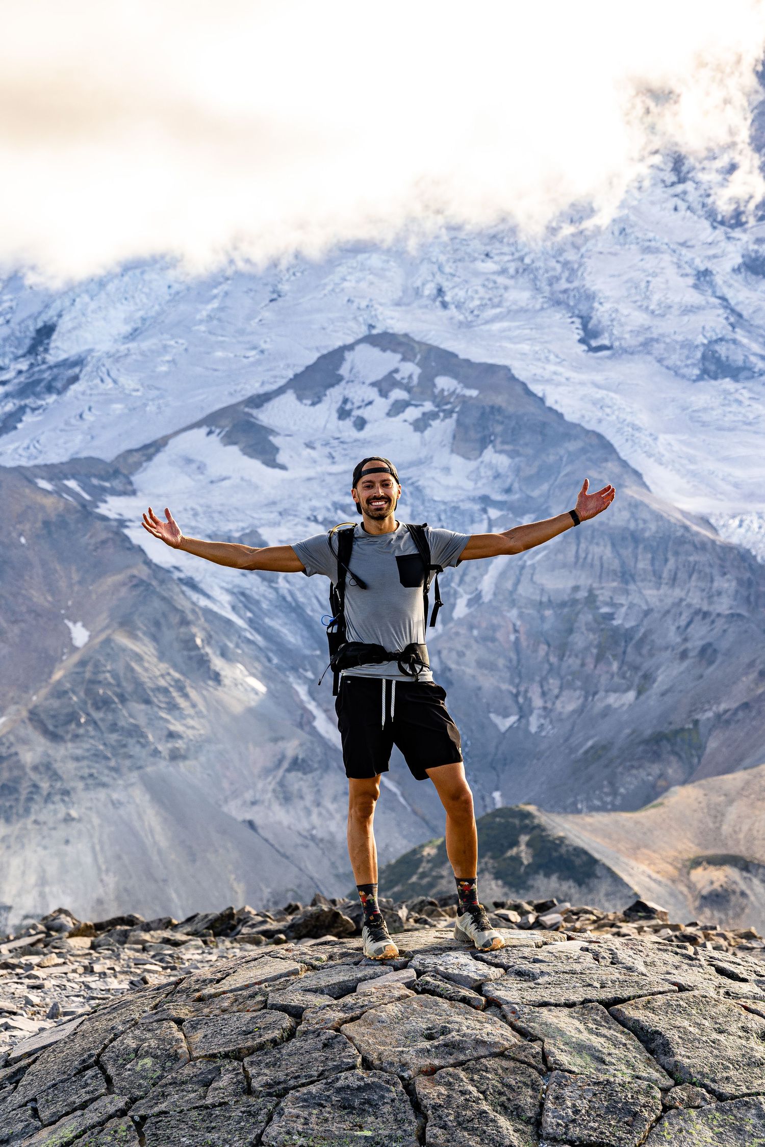 Hiker stands triumphantly with arms outstretched on rocky mountain peak with snow-capped peaks in background.
