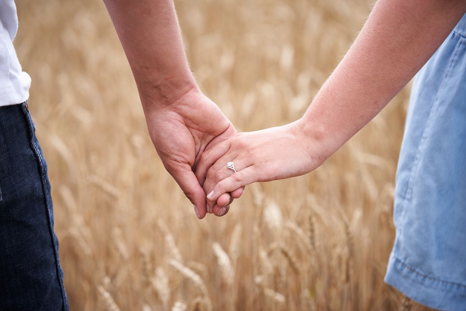 Close-up shot of two people holding hands in a wheat field showcasing an engagement ring.