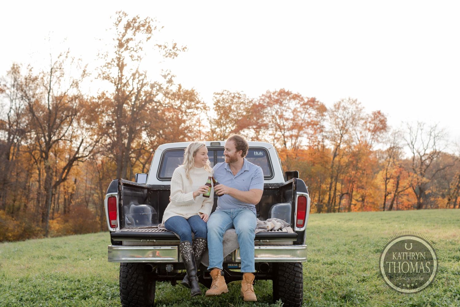 A couple sits together on the tailgate of a vintage pickup truck during sunset in an autumn field.