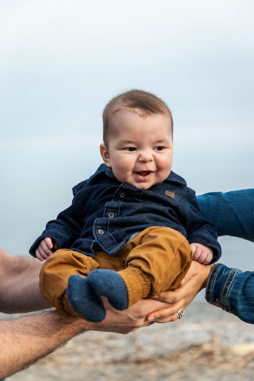 A baby with his mother and fathers hands holding him with the beach in the background
